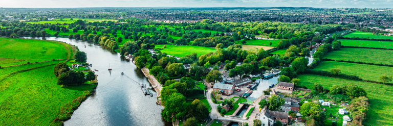 Aerial view of a winding river flowing through a lush green countryside landscape. Trees, fields and open grassland line both banks, with a small waterside settlement, lock and moored boats visible in the foreground. A large town stretches across the horizon beneath a bright blue sky filled with scattered white clouds. The scene highlights the contrast between the river, rural surroundings and urban development in the distance.